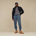 Hombre con chaqueta negra, jersey gris y pantalón jogger azul de punto trenzado, calzando botas chukka UGG Neumel en color chestnut sobre fondo neutro.