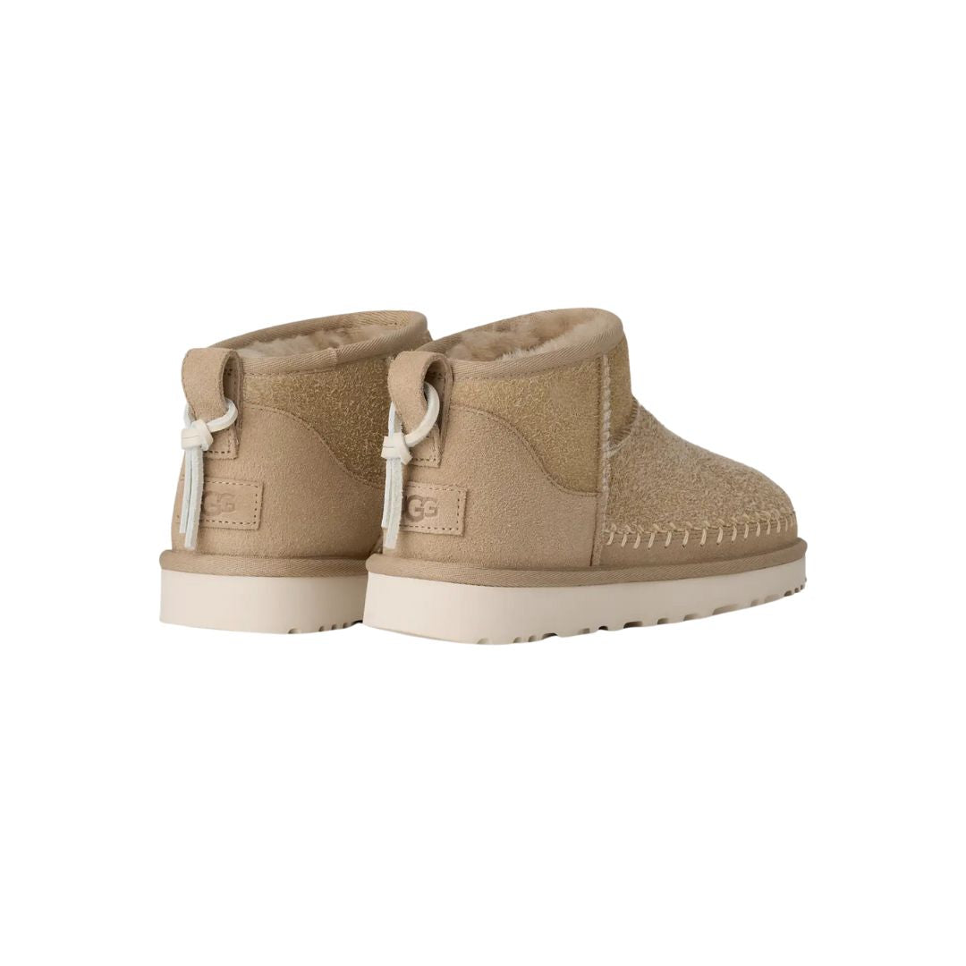 Vista trasera en ángulo de las botas UGG Ultra Mini Biarritz color Mustard Seed con borla decorativa.