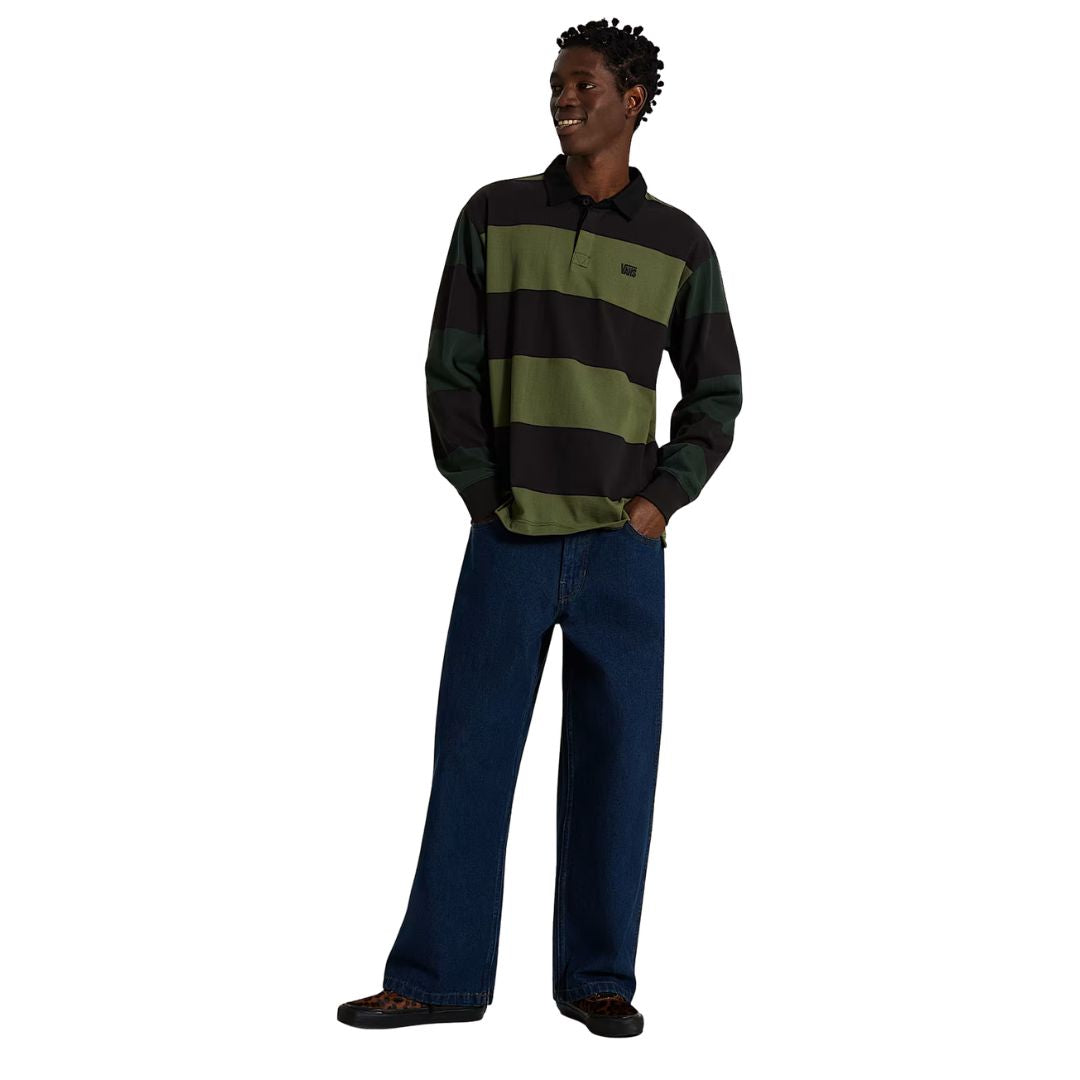 Hombre con polo de rugby Vans Ward de punto a rayas verde y negro, manga larga y cuello negro, combinado con vaqueros anchos azules; vista frontal completa con las manos en los bolsillos.