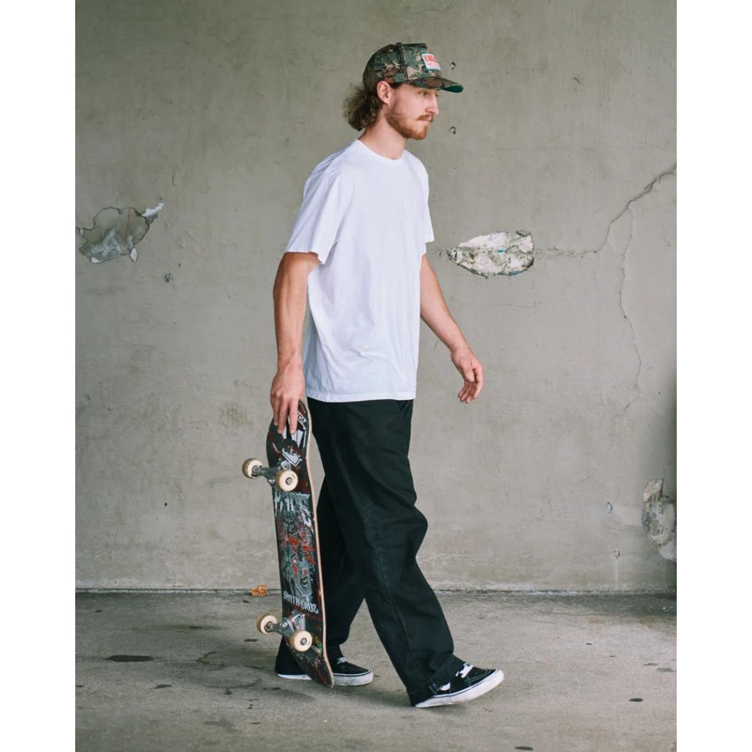 Modelo con vaquero Volcom Billow Denim negro en look urbano skate, gorra camuflaje, camiseta blanca y skate en mano, mostrando la caída ancha del pantalón al caminar.