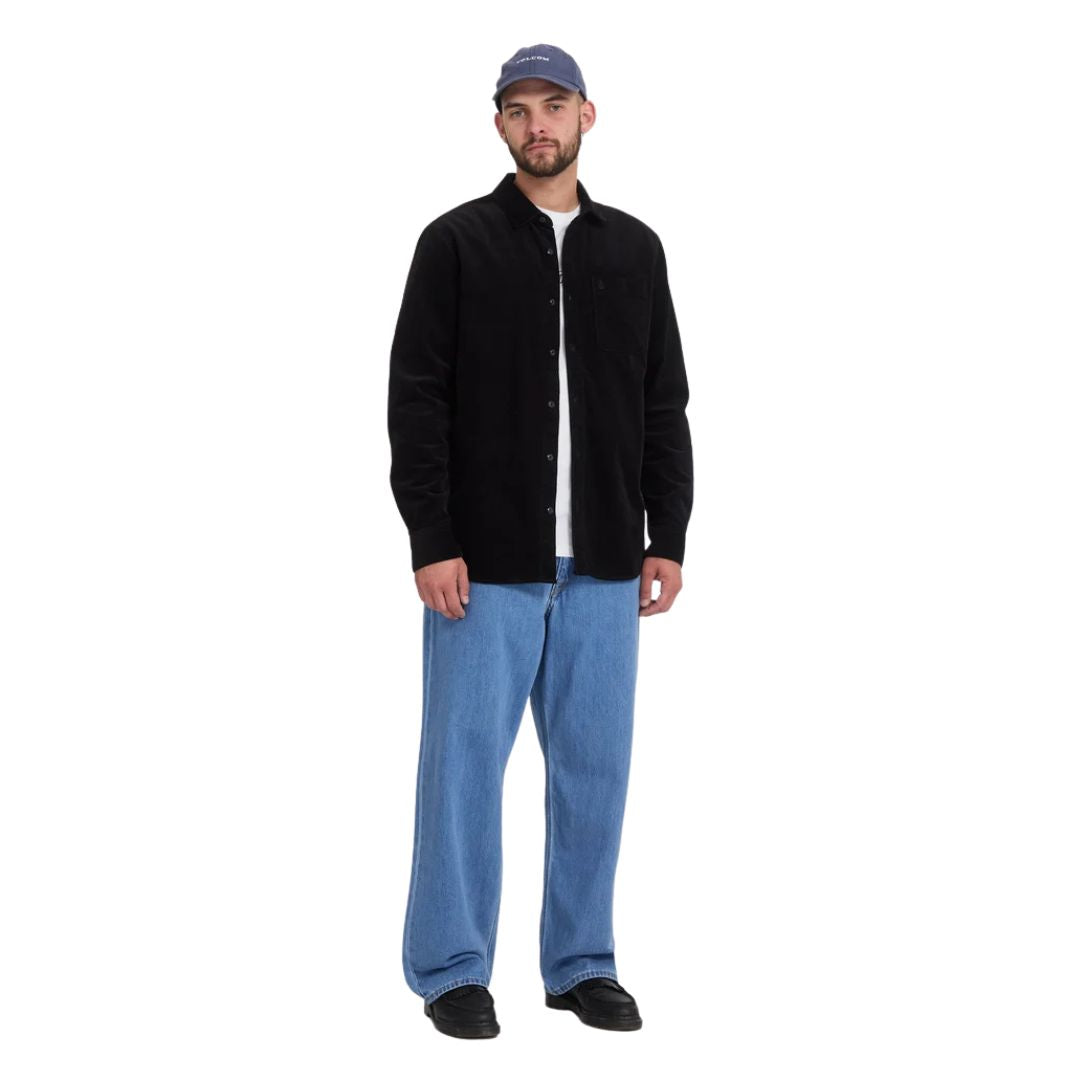 Modelo con pantalón vaquero Volcom Billow Denim en azul Cali Blue, look completo con gorra y camisa negra, mostrando la caída ancha y el tiro bajo del pantalón.