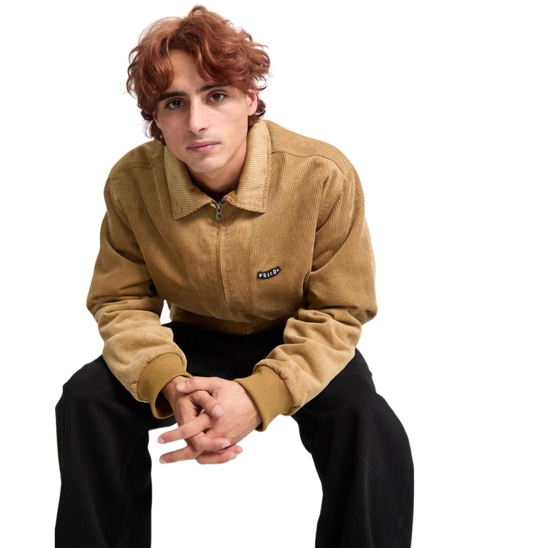 Hombre agachado con chaqueta Volcom Kurtis en color armiño, primer plano frontal con las manos entrelazadas y el logo Volcom bordado en el pecho de la prenda.