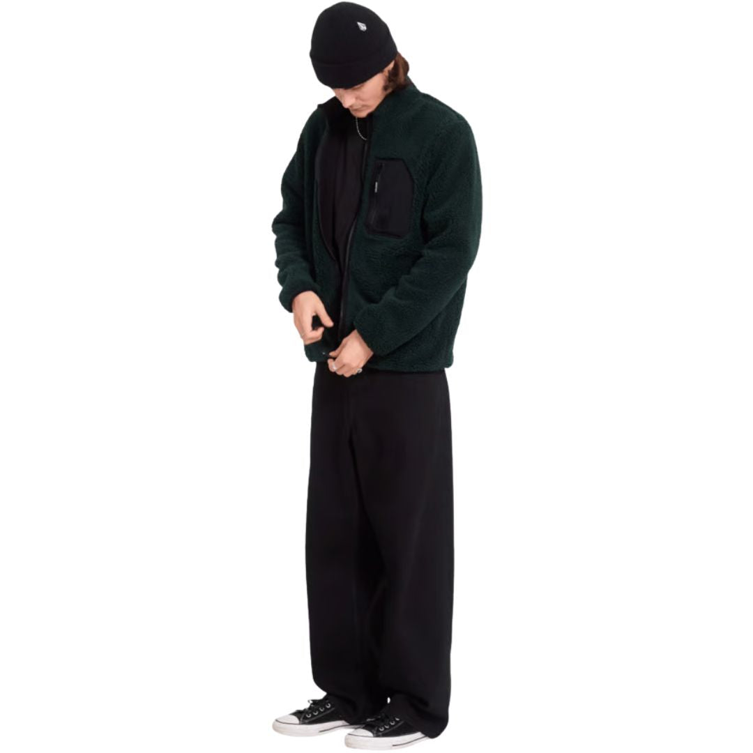 Hombre con sudadera Volcom Muzzer Fuzzar Zip en color verde escarabajo de cuerpo entero, mirando hacia abajo mientras cierra la cremallera del cuello alto.