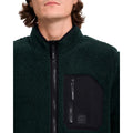 Detalle del bolsillo de pecho con cremallera de la sudadera Volcom Muzzer Fuzzar Zip en color verde escarabajo, con la etiqueta de silicona Volcom Earth Tripper visible.