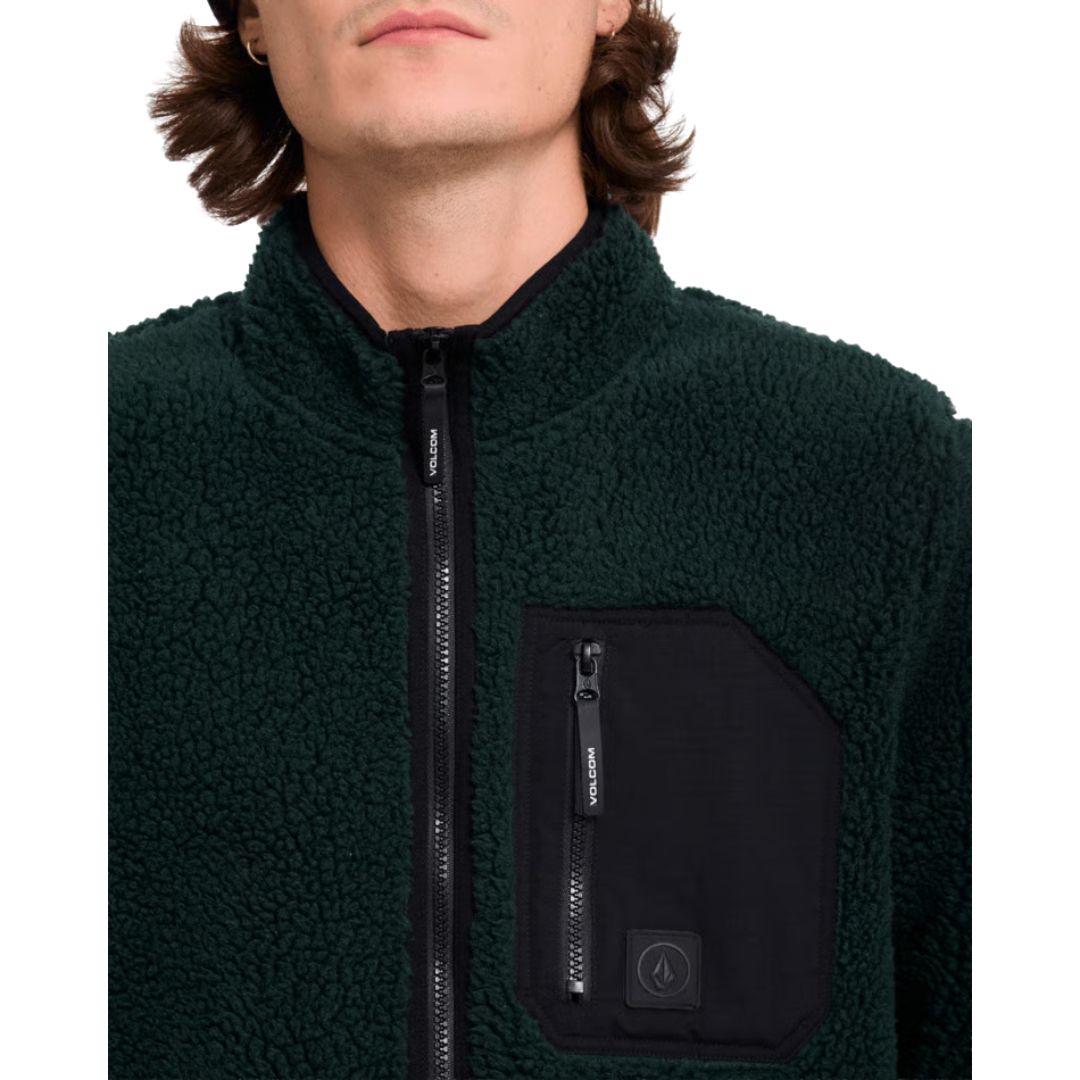 Detalle del bolsillo de pecho con cremallera de la sudadera Volcom Muzzer Fuzzar Zip en color verde escarabajo, con la etiqueta de silicona Volcom Earth Tripper visible.