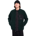Hombre con gorro y sudadera Volcom Muzzer Fuzzar Zip en color verde escarabajo, vista frontal de medio cuerpo con las manos en los bolsillos y la cremallera subida.