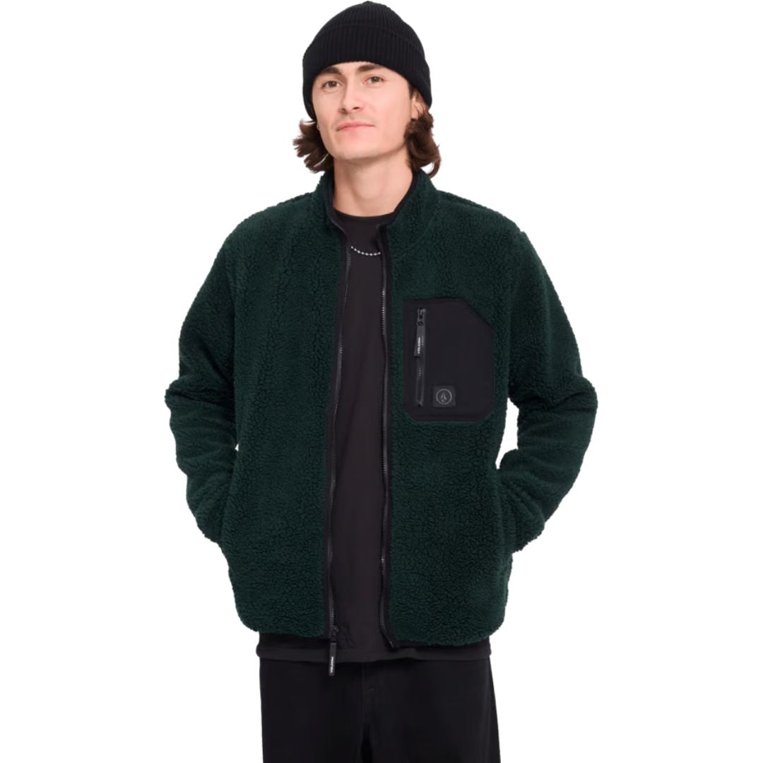 Hombre con gorro y sudadera Volcom Muzzer Fuzzar Zip en color verde escarabajo, vista frontal de medio cuerpo con las manos en los bolsillos y la cremallera subida.