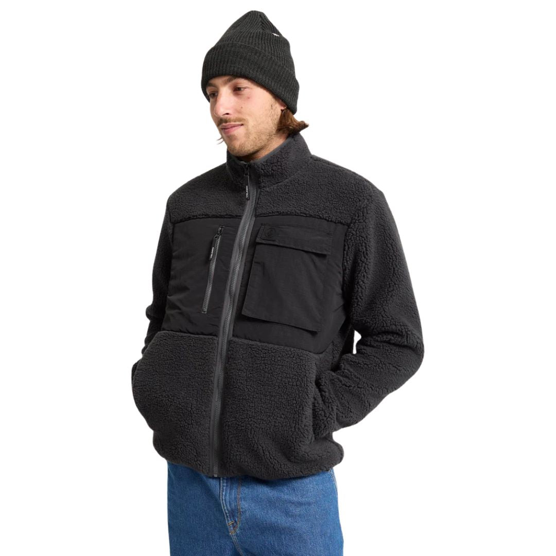 Modelo de pie con chaqueta Volcom Storm Stone Zip negro con las manos en los bolsillos laterales tip canguro.