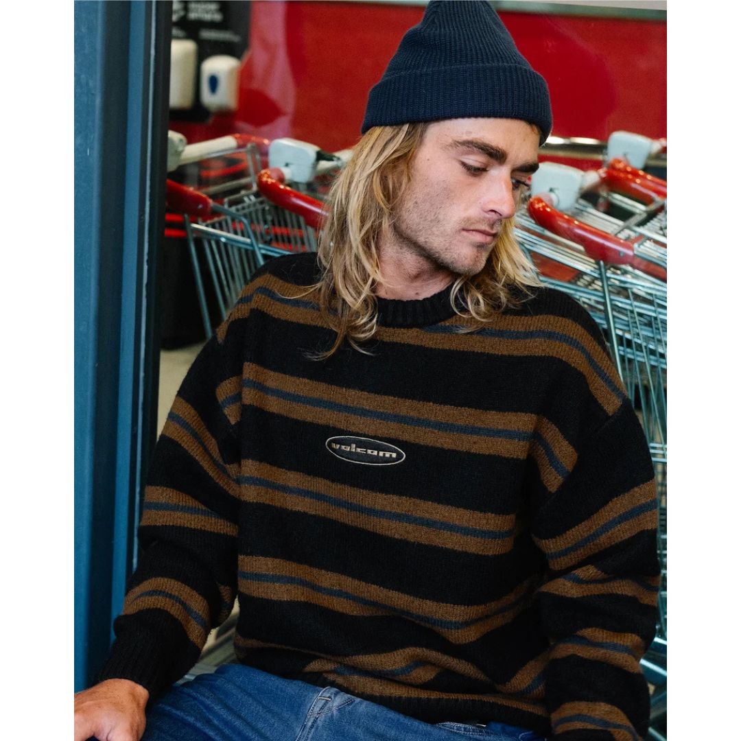 Hombre sentado con gorro y jersey Volcom Striperstand en color negro de rayas, primer plano en escena urbana mostrando el logo Volcom bordado en el pecho.