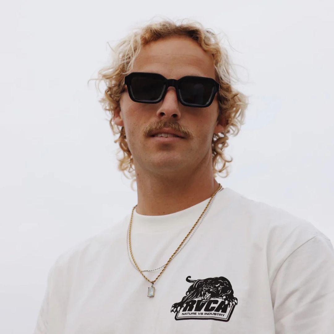 Hombre con gafas de sol Warmouth Dustin Black Black puestas, lente naranja y montura negra, con tabla de surf al fondo.