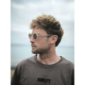 Hombre con gafas de sol Warmouth Grunge Brown puestas de perfil, montura ovalada de acetato camel, fondo de mar y cielo nublado.