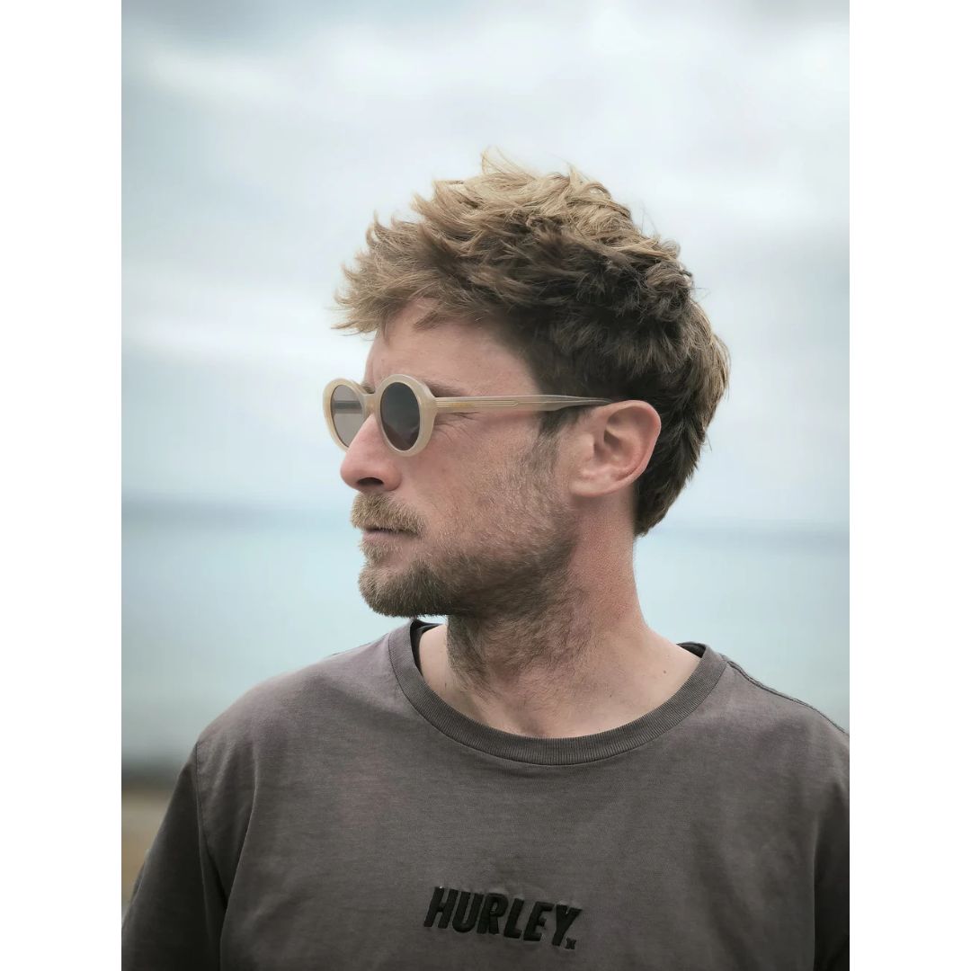 Hombre con gafas de sol Warmouth Grunge Brown puestas de perfil, montura ovalada de acetato camel, fondo de mar y cielo nublado.