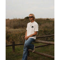 Hombre con gafas de sol Warmouth Iconic Black puestas de perfil, montura envolvente negra, look surfero en entorno natural.